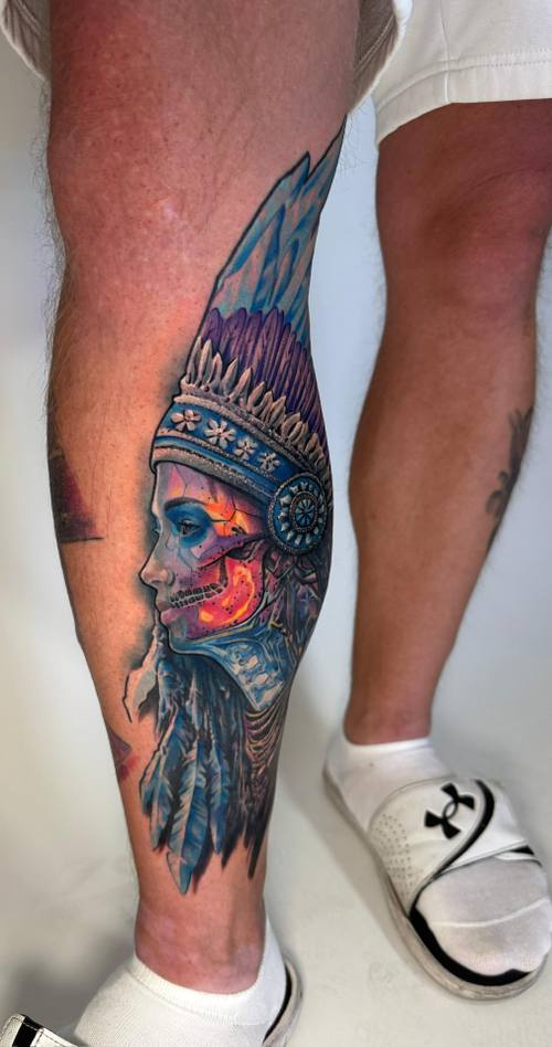 Color realism portrait of an Indigenous figure in a feathered headdress on the lower leg by Padre at KAZENTATTOO, Los Angeles