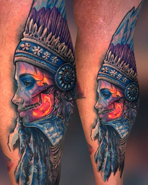 Color realism portrait of an Indigenous woman with a skull face and feathered headdress on the lower leg by Padre at KAZENTATTOO, Los Angeles