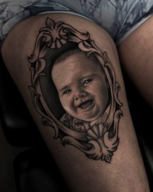 Portrait of a laughing baby in a baroque frame, black and grey thigh tattoo by Sam at KAZENTATTOO, Los Angeles