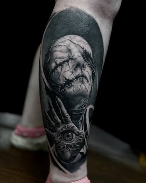 Stitched horror face above a hand holding an eye, black and grey calf tattoo by Sam at KAZENTATTOO, Los Angeles